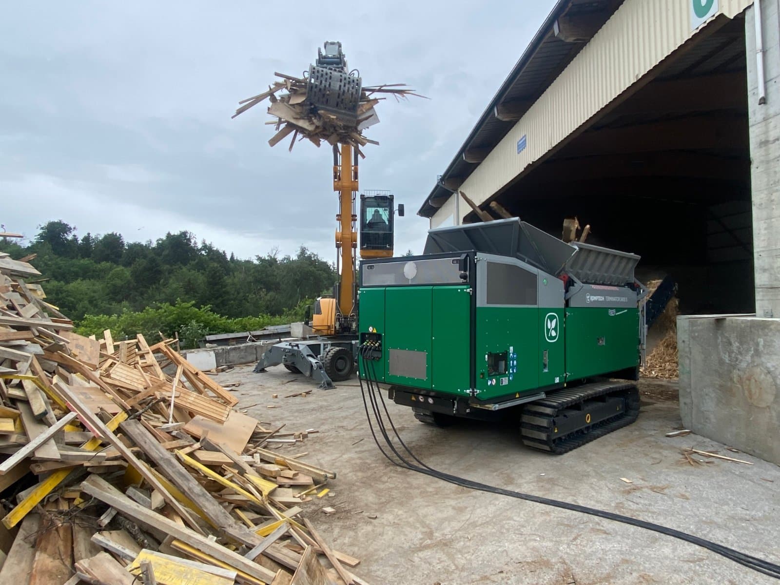 Ein Bagger belädt den vollelektrischen Schredder Komptech TERMINATOR 3400S e-mobile der Société Fribourgeoise de Recyclage (SFR) mit Holzabfällen.